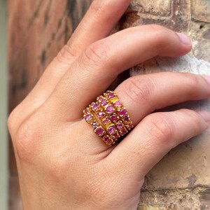 Könnte beinhalten: Ein goldfarbener Ring mit mehreren Reihen kleiner, runder, rosa Edelsteine. Der Ring befindet sich an einem Finger einer Person, wobei die Hand im Vordergrund ist. Der Hintergrund ist eine verschwommene Ansicht einer Backsteinmauer.