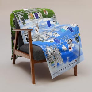 May include: A throw blanket draped over a gray armchair. The blanket features a blue sky and cloud design with various images and text. The blanket is printed with a green grass design on the back side.