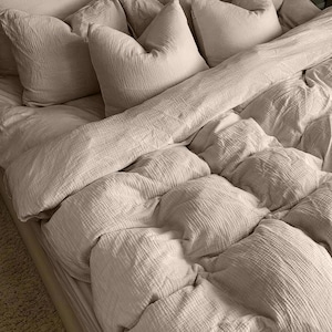 Op de afbeelding: Een bed met beige beddengoed, waaronder verschillende kussens en een dekbed. De kussens zijn aan het hoofdeinde van het bed geplaatst en het dekbed is netjes opgemaakt. Het beddengoed heeft een textuur.