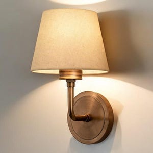 Könnte beinhalten: Eine Wandleuchte mit beigem Lampenschirm und einem Sockel aus bronzefarbenem Metall. Die Lampe hat einen gebogenen Arm und eine kreisförmige Rückplatte. Das Licht wirft einen warmen Schein an die Wand.