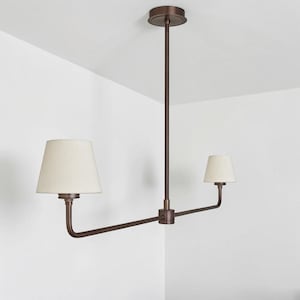 May include: A bronze-colored, two-light pendant light fixture. The fixture has a central vertical rod attached to a ceiling plate, with two horizontal arms extending outwards. Each arm supports a cream-colored lampshade. The fixture is designed for overhead lighting.