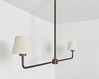 May include: A bronze-colored, two-light pendant light fixture. The fixture has a central vertical rod attached to a ceiling plate, with two horizontal arms extending outwards. Each arm supports a cream-colored lampshade. The fixture is designed for overhead lighting.