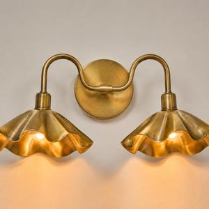 May include: A double wall sconce light fitting in a brushed brass finish. The fitting features a circular wall plate with two curved arms extending outwards, each supporting a wide, scalloped, bell-shaped shade with a warm glowing light.