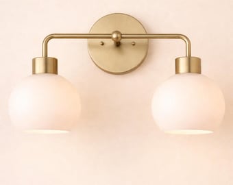 May include: A two-light vanity fixture with a brass finish. The fixture features two spherical, white glass shades and a circular backplate. The light fixture is designed for wall mounting and provides a warm, ambient glow.