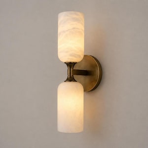 May include: A wall-mounted light fixture featuring a circular antique brass backplate and a horizontal arm supporting two vertical, cylindrical, translucent white glass shades with a marbled texture that emit a soft, warm, diffused glow.