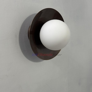 May include: A contemporary wall light with a dark bronze disc and a white globe-shaped lamp. The light is mounted on a light grey wall. The design is minimalist and modern, suitable for various interior styles.