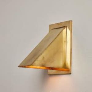 May include: A wall-mounted light fixture featuring a triangular, wedge-shaped brass shade with a brushed metallic finish. The design includes a rectangular backplate and an open bottom that directs light downward, creating a warm glow.