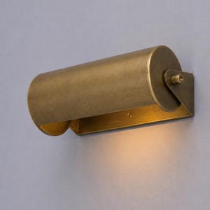 May include: A brass wall light. The cylindrical shade has a curved, open base, and is mounted on a rectangular plate. The light is angled downwards, creating a warm glow. The fixture is designed for wall mounting.