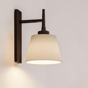 Puede incluir: Aplique de pared con marco de metal marrón oscuro y pantalla de tela beige. El aplique tiene un diseño minimalista, con una base rectangular y un brazo horizontal que sostiene la pantalla. La pantalla es cónica y emite una luz suave y cálida.