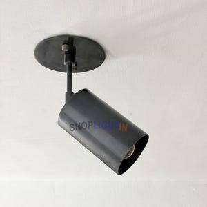 Puede incluir: Un foco negro, cilíndrico, montado en un techo blanco. La luminaria tiene una base circular y un brazo giratorio. El texto "SHOPLIGHTIN" está impreso en el lateral del foco.
