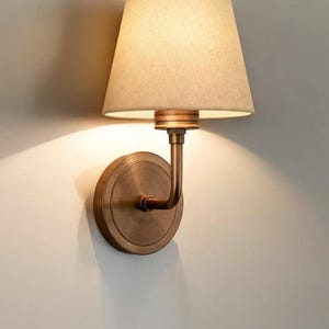 Peut inclure: Une applique murale de couleur bronze avec un abat-jour beige. La lampe a une base circulaire et un bras incurvé supportant l'abat-jour. L'abat-jour conique émet une lumière chaude, projetant des ombres sur le mur.