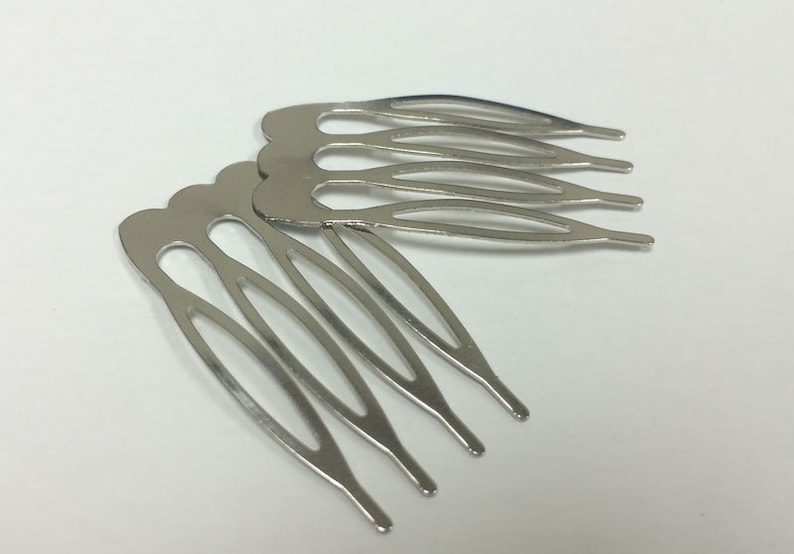 May include: Two silver-colored hair combs with a curved, scalloped top and four tines each. The combs are made of a shiny metal and are lying on a white surface. These hair accessories are designed to be inserted into the hair.