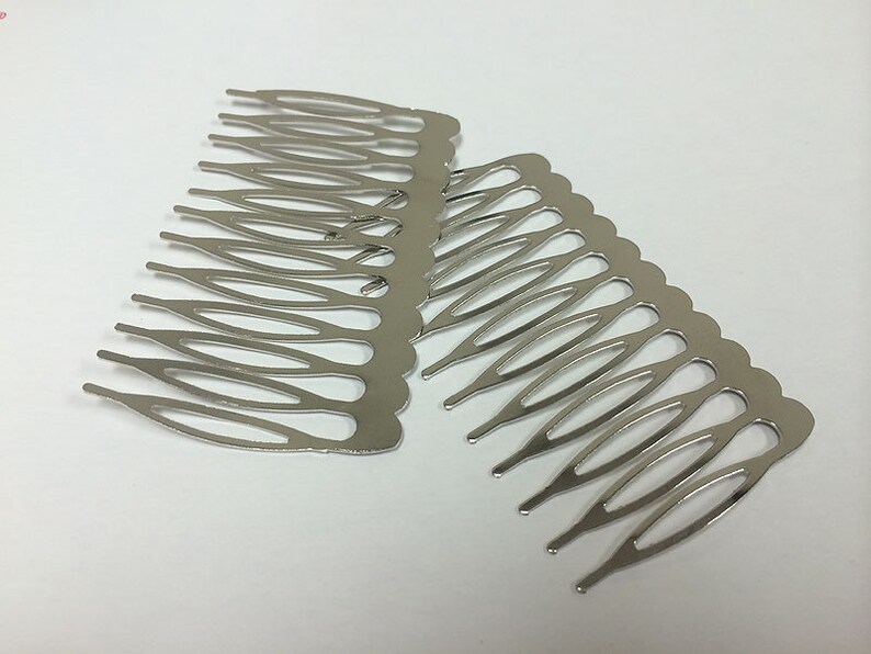 May include: Two silver-colored metal hair combs. Each comb has a series of teeth designed to secure hair. The combs are made of a shiny, reflective metal and are positioned on a white surface.