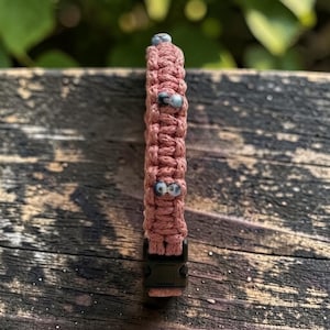 Puede incluir: Una pulsera de paracord color rosa empolvado con una serie de pequeñas cuentas moteadas en gris y blanco tejidas en el centro del cordón. La pulsera se asegura con una hebilla de plástico negro de liberación lateral y muestra un patrón de nudos.