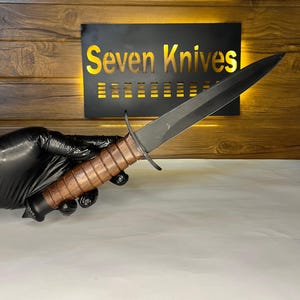 May include: A black dagger with a brown leather-wrapped handle is held by a gloved hand. The blade is dark and pointed. The text "Seven Knives" is displayed on a black sign with a wooden background.
