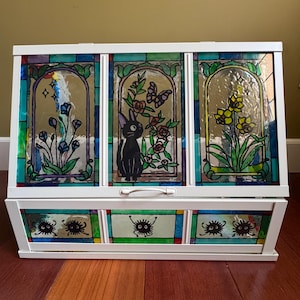 Puede incluir: Cofre blanco con paneles de vidrieras con diseños florales y de gatos. Los paneles superiores representan flores, un gato negro y mariposas. El panel inferior tiene criaturas de dibujos animados. El cofre está sobre un suelo de madera.