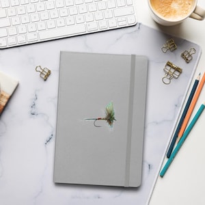 Puede incluir: Un cuaderno gris con una ilustración de una mosca de pesca. El cuaderno está cerrado y tiene un cierre elástico. También se ven un teclado, lápices y una taza de café.