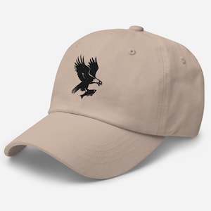 May include: A tan baseball cap featuring a black graphic of an eagle carrying a fish. The cap has a curved brim and a button on top. The design is a classic and simple style.