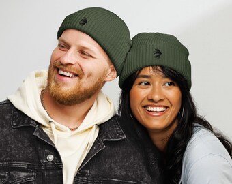 May include: Two people wearing olive green ribbed knit beanies with a black embroidered design. The beanies are the focal point, with the wearers smiling. The background is a plain white.