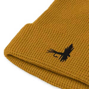 Puede incluir: Gorro de color amarillo mostaza con textura de punto de gofre. Un señuelo de pesca con mosca bordado en negro está en la parte delantera. El gorro está doblado en el borde.