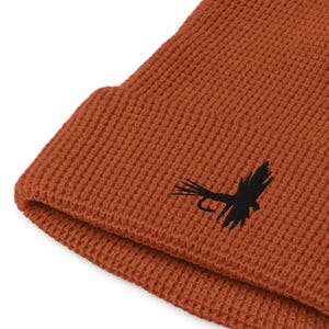 Könnte beinhalten: Eine rostfarbene Beanie-Mütze mit Waffelstrickstruktur. Auf der Vorderseite befindet sich ein schwarzer, gestickter Fliegenfischköder. Die Mütze ist am Rand gefaltet.