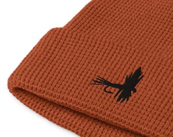 May include: A rust-colored beanie hat with a waffle knit texture. A black embroidered fly fishing lure is on the front. The hat is folded at the brim.