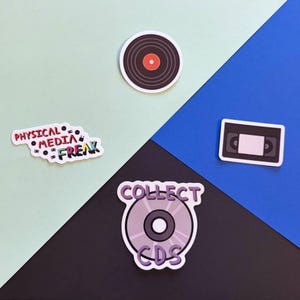 以下が含まれることがあります： 赤い中心を持つ黒いレコード、黒いVHSテープ、COLLECT CDSという文字が入った薄紫色のCD、そして小さなメディアアイコンに囲まれたカラフルなPHYSICAL MEDIA FREAKという文字が入った白いステッカーの4枚のステッカー。