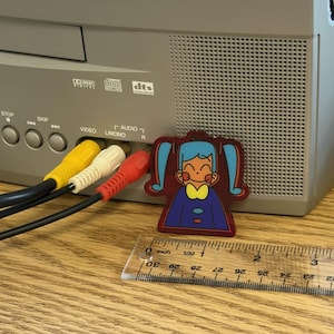 May include: A colorful cartoon-style pin featuring a girl with blue pigtails, a yellow bow, and a blue dress. The pin is next to a ruler showing inch measurements. The pin is approximately 1 inch in height. The background includes a gray electronic device with audio and video input jacks.