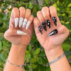 May include: Two hands displaying long, pointed false nails. One hand features white nails with silver designs and rhinestones. The other hand showcases black nails with silver accents and rhinestones. The nails are a fashionable accessory.