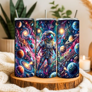 Peut inclure: Un gobelet cylindrique avec une paille en métal arborant un imprimé spatial vibrant. Le motif représente un astronaute central flottant au milieu de nébuleuses colorées, de planètes, d'étoiles et de vaisseaux spatiaux sur un fond cosmique bleu et violet.