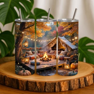 Puede incluir: Tres vasos aislados de acero inoxidable con un diseño temático de otoño. El diseño presenta una escena al aire libre acogedora con una fogata, una tienda de campaña, calabazas y follaje otoñal. Cada vaso tiene una pajita y tapa plateadas.