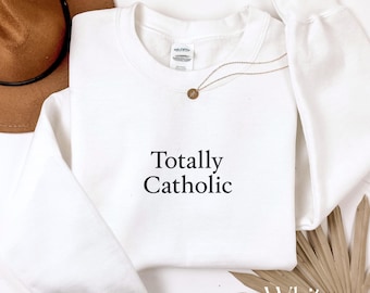 Puede incluir: Sudadera blanca de cuello redondo con el texto "Totally Catholic" en negro. Un collar dorado con un pequeño colgante descansa sobre la camisa. La palabra "White" está en la esquina inferior derecha de la imagen.