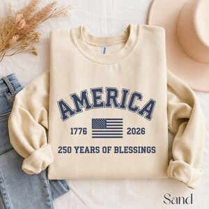 Pu&ograve; includere: Felpa color sabbia con la scritta "AMERICA" in lettere blu navy ad arco. Sotto, la bandiera americana, i numeri "1776" e "2026" e il testo "250 YEARS OF BLESSINGS".