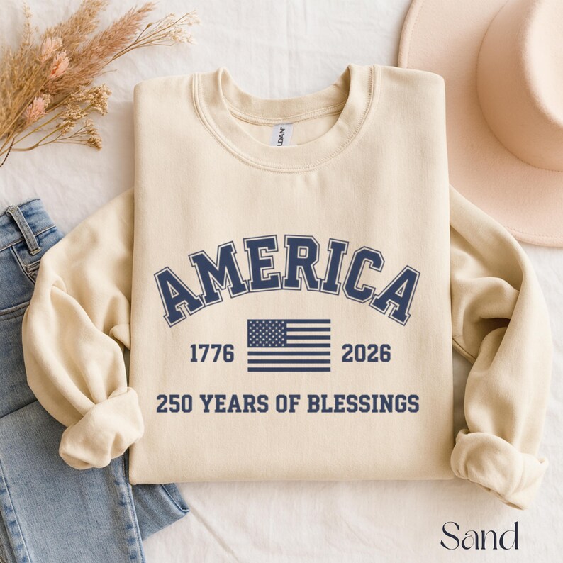 Pu&ograve; includere: Felpa color sabbia con la scritta "AMERICA" in lettere blu navy ad arco. Sotto, la bandiera americana, i numeri "1776" e "2026" e il testo "250 YEARS OF BLESSINGS".