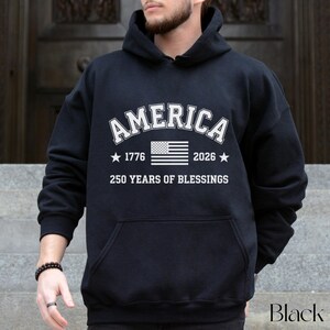 Pu&ograve; includere: Felpa con cappuccio nera con tasca frontale. Il testo bianco sul davanti recita "AMERICA" sopra una grafica della bandiera americana, con "1776" e "2026" su entrambi i lati. Sotto la bandiera, c'&egrave; scritto "250 YEARS OF BLESSINGS".