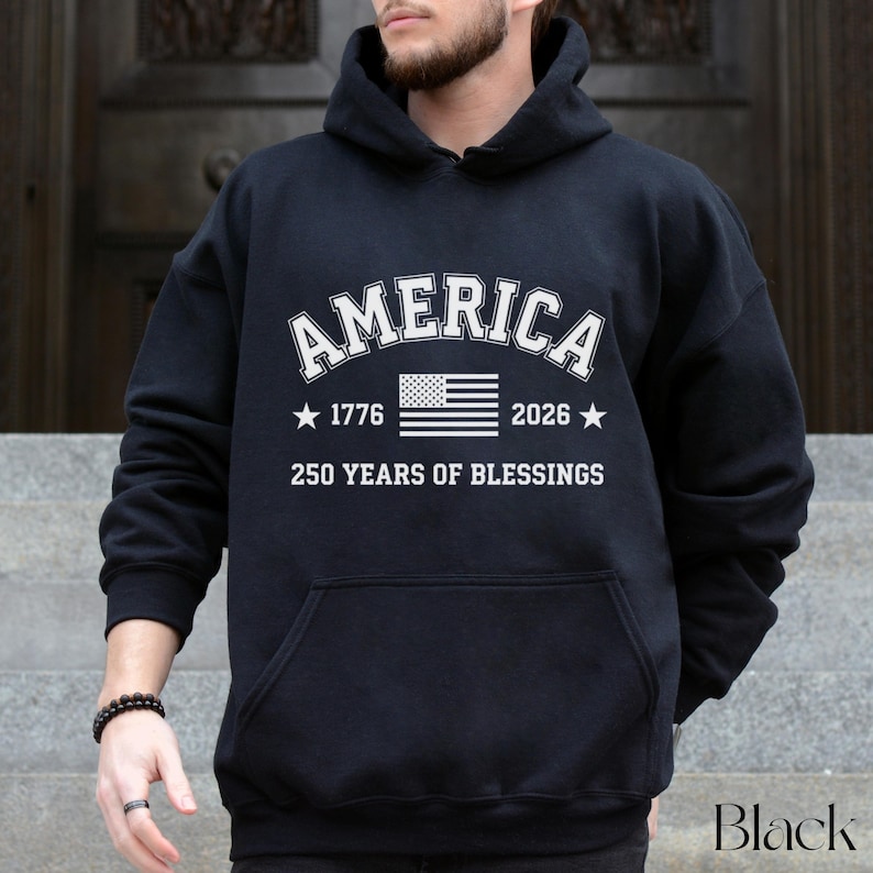 Pu&ograve; includere: Felpa con cappuccio nera con tasca frontale. Il testo bianco sul davanti recita "AMERICA" sopra una grafica della bandiera americana, con "1776" e "2026" su entrambi i lati. Sotto la bandiera, c'&egrave; scritto "250 YEARS OF BLESSINGS".
