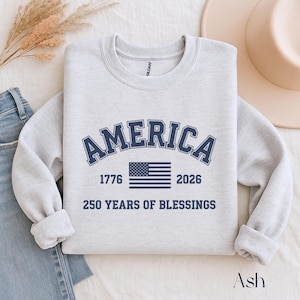 Pu&ograve; includere: Una felpa grigio chiaro con la scritta "AMERICA" in lettere arcuate blu navy sopra una bandiera americana blu navy. Sotto la bandiera ci sono i numeri "1776" e "2026". Il testo "250 YEARS OF BLESSINGS" &egrave; sotto.