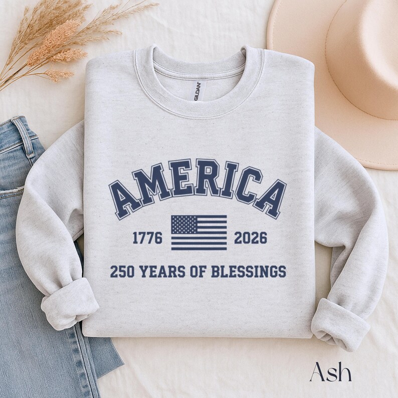Pu&ograve; includere: Una felpa grigio chiaro con la scritta "AMERICA" in lettere arcuate blu navy sopra una bandiera americana blu navy. Sotto la bandiera ci sono i numeri "1776" e "2026". Il testo "250 YEARS OF BLESSINGS" &egrave; sotto.