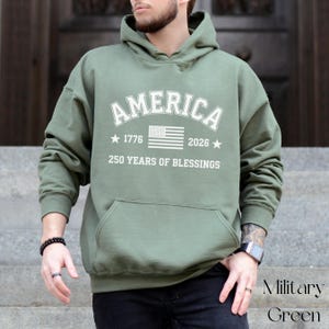 Pu&ograve; includere: Felpa con cappuccio verde militare con la scritta "AMERICA" in bianco. Il design include la bandiera americana, gli anni 1776 e 2026 e la frase "250 YEARS OF BLESSINGS". La felpa ha una tasca frontale.