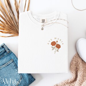 Peut inclure: T-shirt blanc avec l'inscription "AVE MARIA" et un motif floral orange et beige. Un collier argent&eacute; et un jean bleu sont &eacute;galement visibles. L'&eacute;tiquette de la marque "COMFORT COLORS" est visible sur le t-shirt.