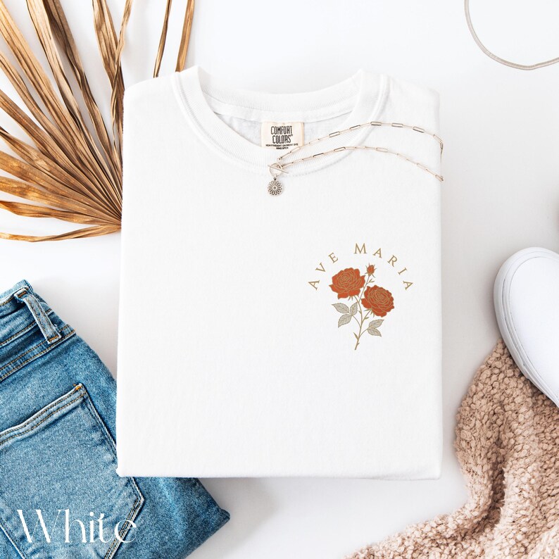 Peut inclure: T-shirt blanc avec l'inscription "AVE MARIA" et un motif floral orange et beige. Un collier argent&eacute; et un jean bleu sont &eacute;galement visibles. L'&eacute;tiquette de la marque "COMFORT COLORS" est visible sur le t-shirt.