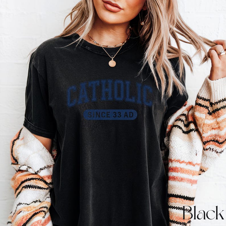 May include: Black t-shirt with the word "CATHOLIC" in blue block letters above "SINCE 33 AD". The shirt is worn with a patterned sweater. The word "Black" is printed in the lower right corner.