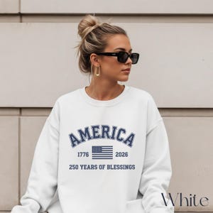 Pu&ograve; includere: Felpa bianca con la scritta "AMERICA" in testo blu navy arcuato, insieme agli anni "1776" e "2026" e una grafica della bandiera americana. Il testo "250 YEARS OF BLESSINGS" &egrave; sotto la bandiera. La parola "White" &egrave; nell'angolo in basso a destra.