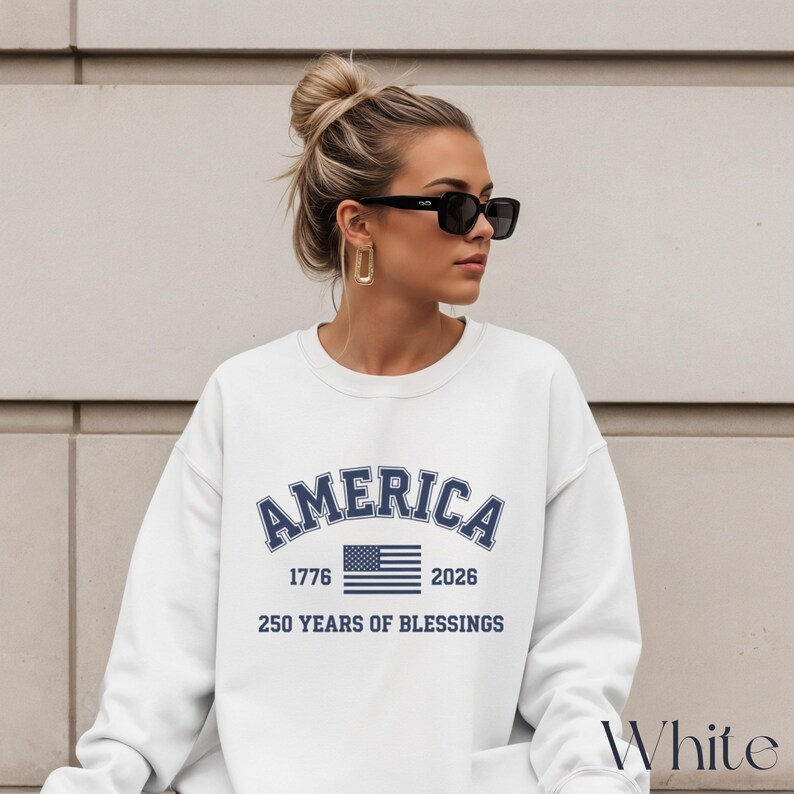 Pu&ograve; includere: Felpa bianca con la scritta "AMERICA" in testo blu navy arcuato, insieme agli anni "1776" e "2026" e una grafica della bandiera americana. Il testo "250 YEARS OF BLESSINGS" &egrave; sotto la bandiera. La parola "White" &egrave; nell'angolo in basso a destra.