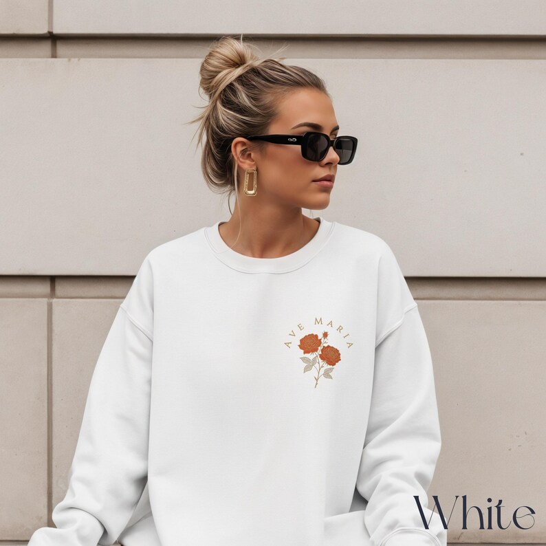 Peut inclure: Sweat-shirt blanc avec l'inscription "Ave Maria" et un motif floral au centre. Le mot "White" est &eacute;crit en bas &agrave; droite. Le sweat-shirt est de style col rond.