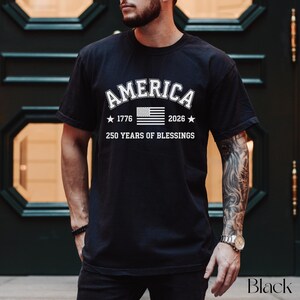 Pu&ograve; includere: T-shirt nero con la scritta "AMERICA" in lettere bianche arcuate, una grafica della bandiera americana, gli anni "1776" e "2026" con stelle e la scritta "250 YEARS OF BLESSINGS".