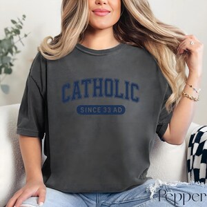 May include: Dark gray t-shirt with the word "CATHOLIC" in navy blue arched lettering above "SINCE 33 AD" in a navy blue rectangular box. The shirt has a slightly faded, vintage look.