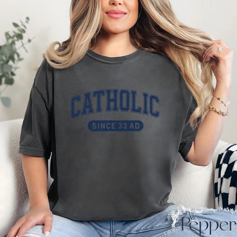 May include: Dark gray t-shirt with the word "CATHOLIC" in navy blue arched lettering above "SINCE 33 AD" in a navy blue rectangular box. The shirt has a slightly faded, vintage look.