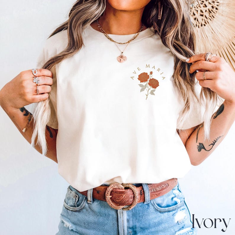 Peut inclure: T-shirt ivoire avec l'inscription "AVE MARIA" au-dessus d'un motif floral de deux roses couleur rouille. Le t-shirt est de couleur neutre et a un col rond classique. Le mot "Ivory" est imprim&eacute; en bas &agrave; droite du t-shirt.