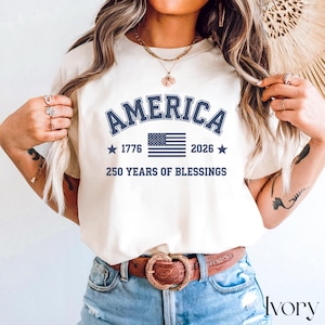 Pu&ograve; includere: T-shirt color crema con la scritta "AMERICA" in blu navy nella parte superiore, con "1776" e "2026" ai lati di una bandiera americana blu navy. Il testo "250 YEARS OF BLESSINGS" &egrave; sotto la bandiera.